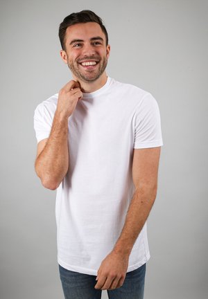 Lachende man met kort donker haar die een effen wit T-shirt en blauwe spijkerbroek draagt tegen een neutrale grijze achtergrond.