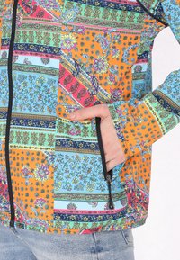 Giacca zip con motivo colorato che presenta sezioni blu, arancioni e rosa con design floreali e geometrici. Include una tasca frontale e dettagli neri.