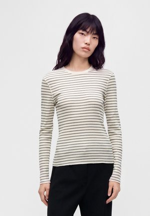 Jeune femme aux longs cheveux noirs portant un haut blanc à manches longues avec de fines rayures horizontales noires et un pantalon noir.