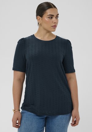 Kobieta o ciemnych włosach związanych w niski kok, ubrana w granatowy, teksturowany top z krótkim rękawem i jasnoniebieskie dżinsy, patrząca na bok na jednolitym tle.