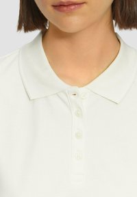 T-shirt polo blanc avec un col classique, patte de boutonnage à cinq boutons et un tissu texturé. Présente un léger détail de couture près du col.