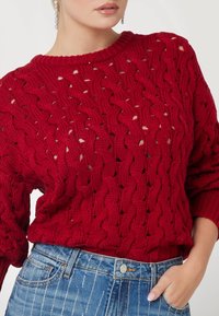 Pull en laine rouge avec un motif en relief, col rond et coupe ample. Associé à un jean en denim à rayures bleues.