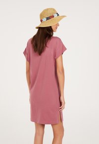 Rosa Baumwollkleid mit kurzen Ärmeln und seitlichen Schlitzen. Kombiniert mit einem geflochtenen Strohhut, der ein buntes Band hat.