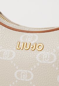 Bolso de mano de cuero texturizado beige con un acento marrón. Presenta el logotipo dorado "LIU·JO" y sutiles patrones blancos en toda la superficie.