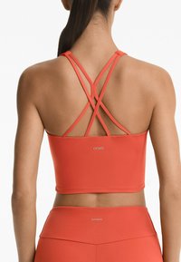 Koralni sportski crop top s križnim trakama na leđima, napravljen od glatkog materijala, s malim logotipom na rubu.