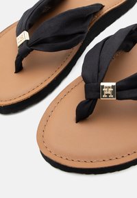 Sandales plates marron avec une sangle en tissu noir nouée et un accent logo métallique, dotées d'une semelle intérieure en cuir lisse et d'une semelle extérieure texturée noire.