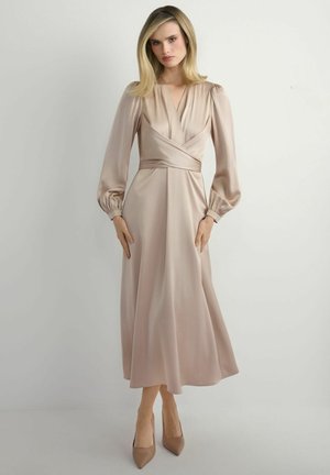 Kobieta ubrana w beżową satynową sukienkę midi z długimi bufiastymi rękawami, dekoltem w kształcie koperty w kształcie litery V, dopasowaną talią i butami na obcasie z ostrym noskiem, stojąca na jednolitym tle.
