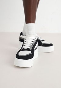 Baskets noir et blanc avec une épaisse semelle blanche, des accents en mesh et en suède, et des détails de panneaux contrastants. Des lacets blancs complètent le design.