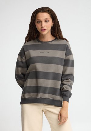 Joven mujer con cabello rizado lleva una sudadera gris a rayas con el texto "Polo Club" y pantalones beige claros sobre un fondo blanco.
