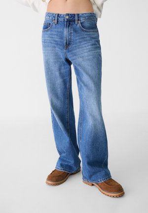 Jean en denim bleu à jambes larges avec un lavage décoloré, comprenant des poches avant, des passants de ceinture et une fermeture à bouton. Stylisé avec des chaussures marron.