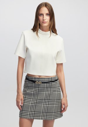 Witte cropped top met korte mouwen en een hoge kraag, gecombineerd met een zwart-wit geruite rok met een zwarte riem en gouden accenten.