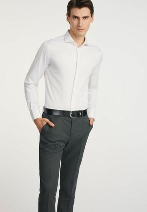 Mężczyzna w białej koszuli i ciemnoszarych spodniach z czarnym paskiem, stojący z rękami w kieszeniach na jednolitym białym tle.