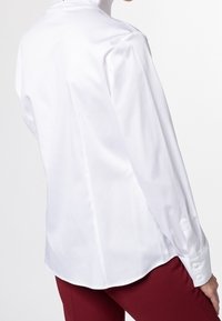 Vit långärmad skjorta med knappar, tillverkad av ett slätt tyg, med hög krage och rundad nederkant, kombinerad med röda byxor.