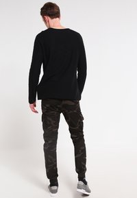 Mężczyzna w czarnej koszuli z długim rękawem, ciemnych spódnicznych spodniach w kamuflaż, oraz szarych sneakersach, stoi tyłem na jednolitym białym tle.