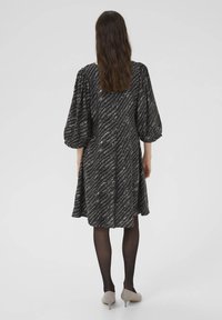 Schwarzes Kleid mit einem wirbelnden metallischen Muster, Puffärmeln und einer ausgestellten Silhouette. Getragen mit schwarzen Strumpfhosen und beigen Absätzen.