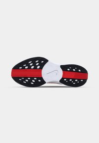 La semelle de la chaussure de sport Nike présente une base en caoutchouc noir avec des accents rouges et une semelle intermédiaire blanche. Texturée avec des motifs circulaires et rectangulaires pour une meilleure adhérence.