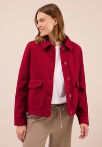 Femme portant une veste boutonnée texturée rouge vif sur une chemise blanche et un pantalon beige à cordon, regardant sur le côté et souriant.
