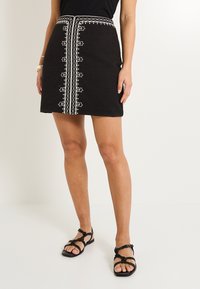 Falda negra con bordado blanco en el centro, que cuenta con una tela texturizada, una forma ajustada y una cinturilla con costuras decorativas.