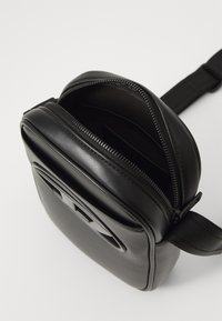 Sac bandoulière en cuir noir avec une fermeture éclair partiellement ouverte et une sangle en tissu ajustable, montrant un intérieur vide et un détail de logo à l'avant.
