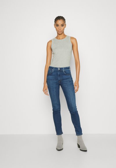 Siva brezrokavnik v kombinaciji z temno modrimi skinny jeans hlačami. Manekenka nosi sive gležnjarje. Ozadje je jasno belo.