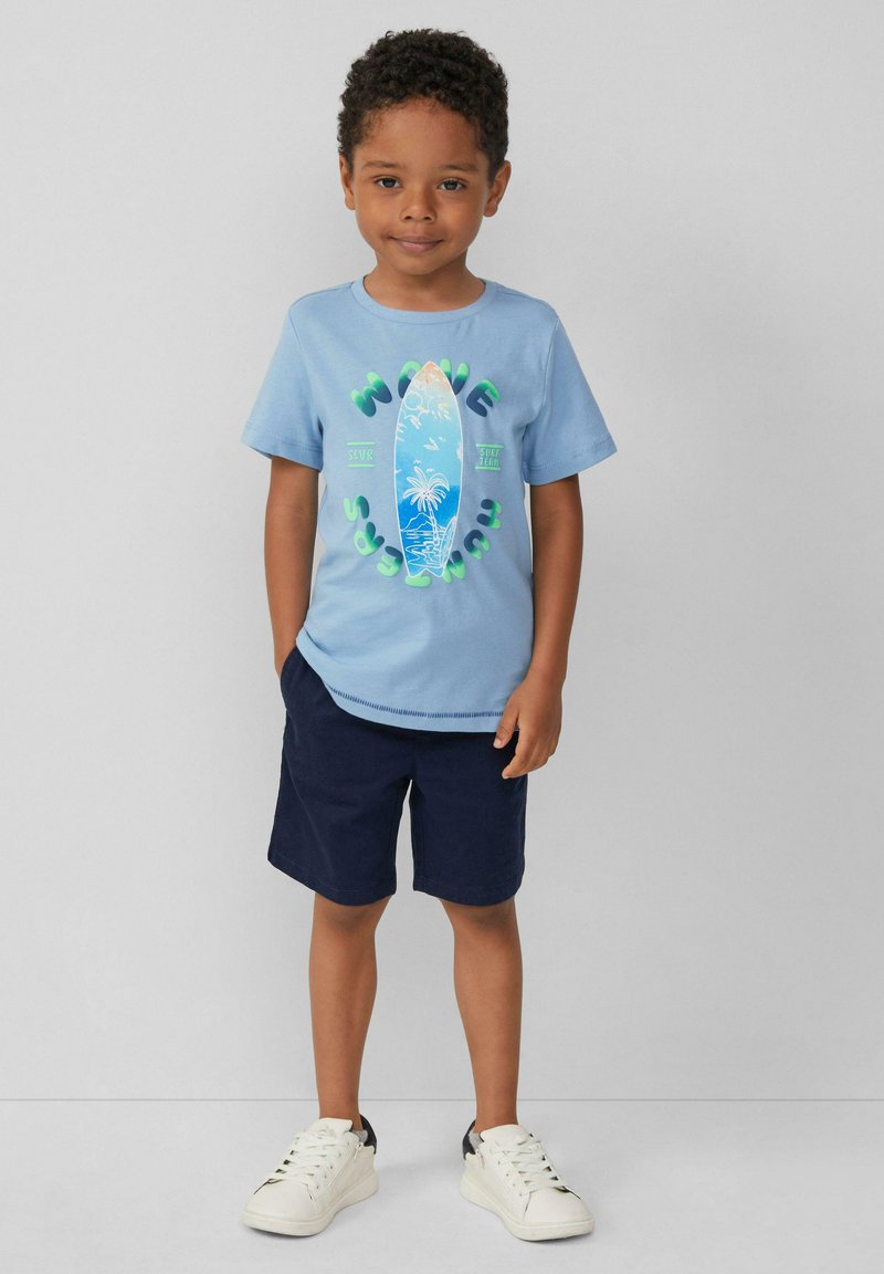 Hellblaue Baumwoll-T-Shirt mit einem Surfbrett-Graphic und Palmen, kombiniert mit marineblauen Shorts und weißen Sneakers. Einfaches und lässiges Design.