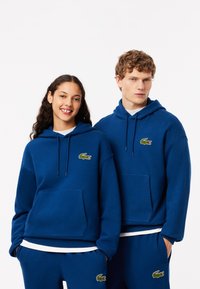 Felpe con cappuccio blu realizzate in morbido tessuto, dotate di una tasca a marsupio e logo di coccodrillo ricamato sul petto.