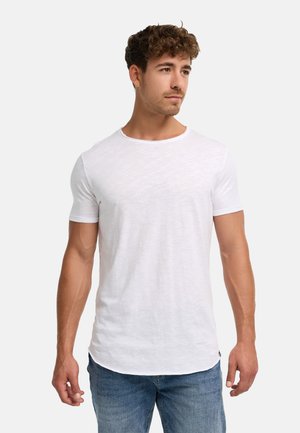 Homme aux cheveux bruns bouclés portant un t-shirt blanc uni à manches courtes et un jean bleu, debout et regardant vers la droite.