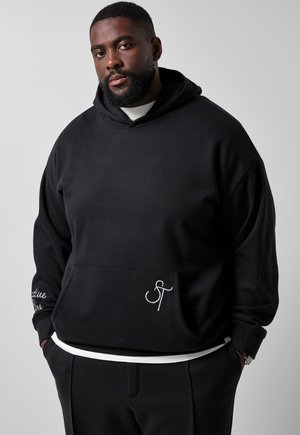 Man med skägg, iklädd svart hoodie och svarta byxor, står mot grå bakgrund med händerna i fickorna.