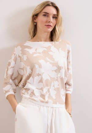 Blonde vrouw draagt een beige-witte trui met bloemenpatroon en driekwart mouwen en witte, hooggetailleerde broek, staand met handen in de zakken.