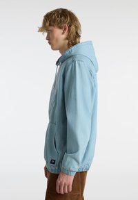 Lichtblauwe denimjas met capuchon, elastische manchetten en een voorzak. Soepele textuur en losse pasvorm, met een koordsluiting.