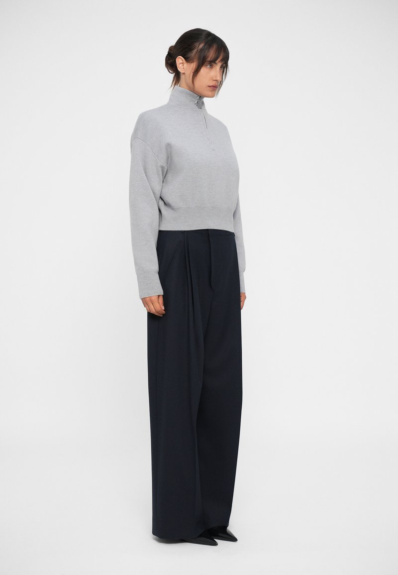 Un pull court zippé gris associé à un pantalon large taille haute bleu marine. Textures lisses, design minimal et détails de plis subtils.