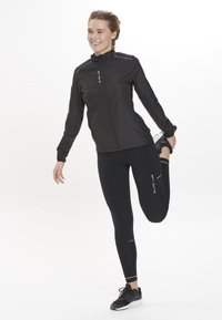 Sort sportjakke med lynlås, refleksdetaljer; parret med sorte leggings med hvid søm. Modellen strækker benet, mens hun har sneakers på.