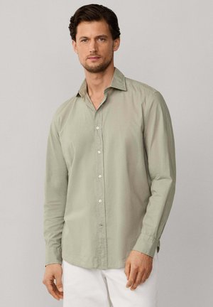 Homme portant une chemise à manches longues vert clair boutonnée et un pantalon blanc, debout devant un fond uni gris clair.