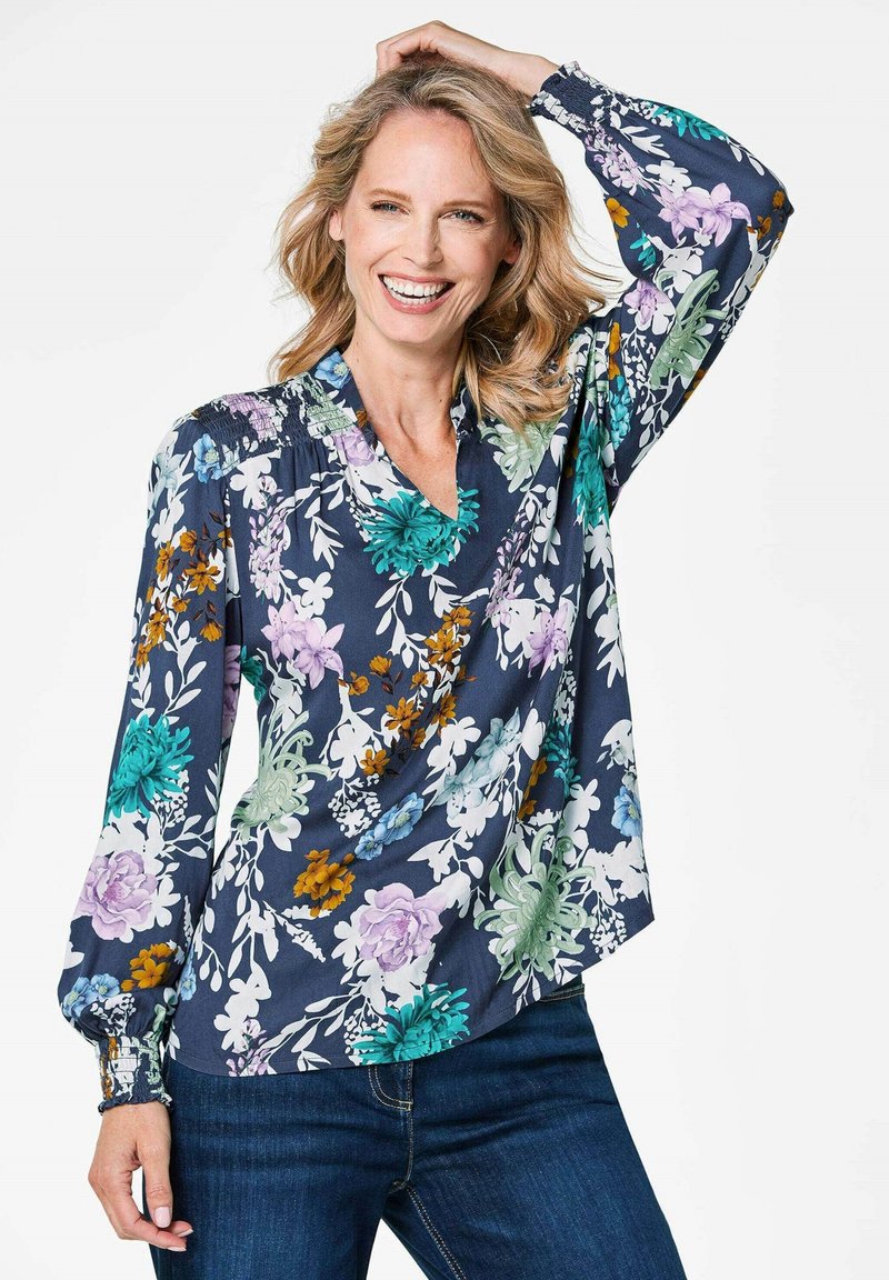 Blumenbluse mit navy Hintergrund, die lebendige Blumen in Grün, Lila und Orange zeigt. Lange Ärmel, V-Ausschnitt und geraffte Details.