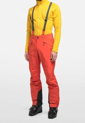 Personne portant un pantalon de ski orange vif avec des bretelles noires, une veste polaire jaune zippée et des chaussures de ski noires.