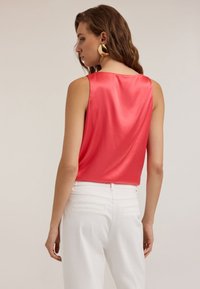 Top sin mangas de satén color coral con un diseño de espalda drapeada, combinado con pantalones blancos de cintura alta y pendientes de aro dorados.