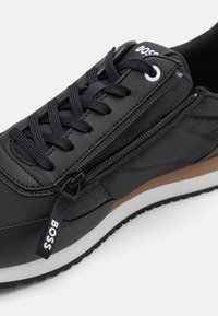 Zapatilla negra que combina materiales lisos y texturizados, con cierre de cremallera, suela marrón contrastante y diseño con cordones.