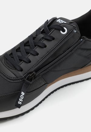 Scarpa da ginnastica nera con una combinazione di materiali lisci e strutturati, chiusura con zip, suola marrone a contrasto e design con lacci.