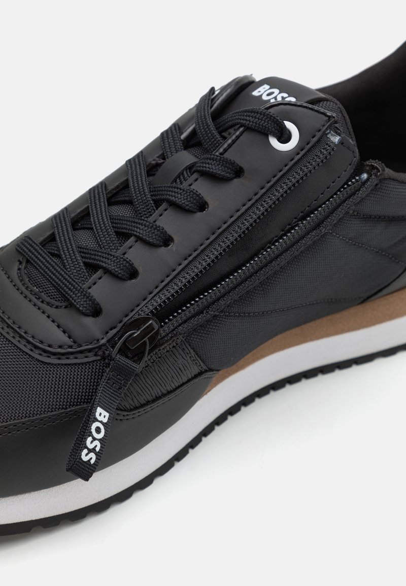 Zapatilla negra que combina materiales lisos y texturizados, con cierre de cremallera, suela marrón contrastante y diseño con cordones.