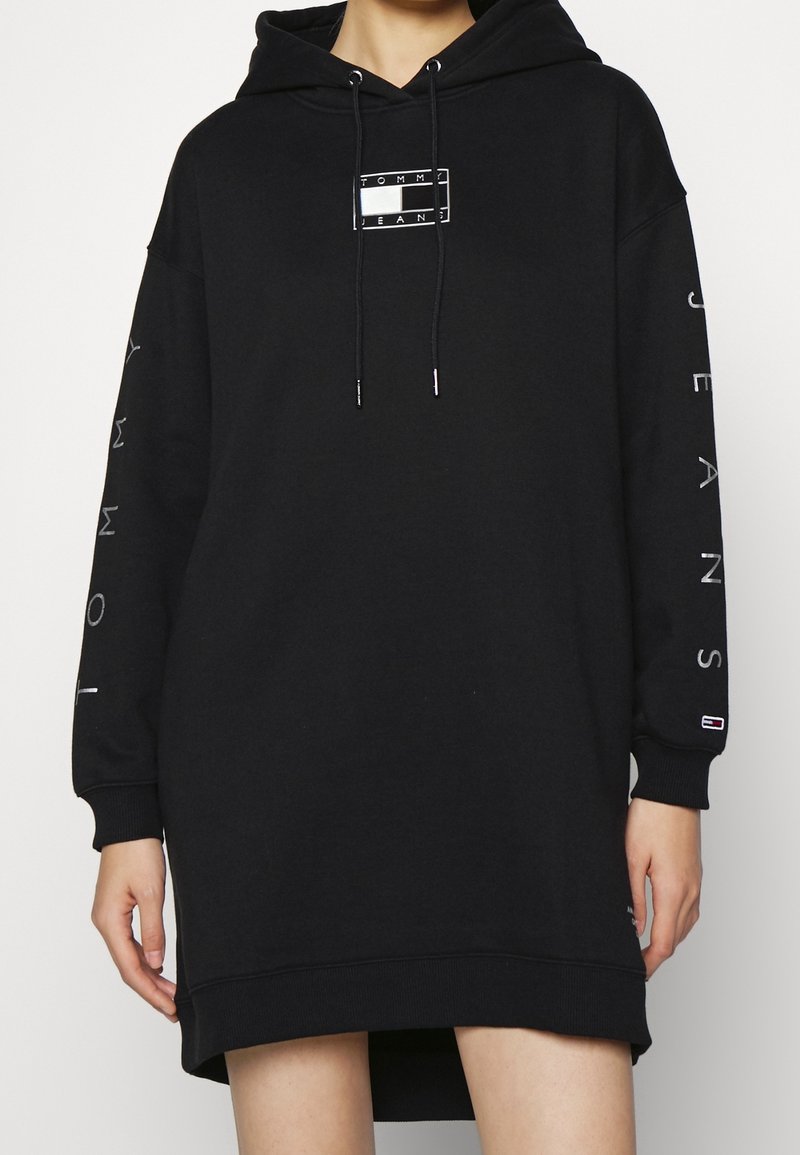 Personne portant une robe sweat à capuche noire oversize avec un logo sur le devant et le texte "Tommy Jeans" sur les manches, debout devant un fond uni.