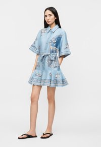 Νέα γυναίκα που φοράει ένα κοντό φόρεμα σε γαλάζιο χρώμα με λουλουδένιο μοτίβο και ζώνη στη μέση, καθώς και μαύρα σαγιονάρα, στέκεται με τα χέρια στις τσέπες.