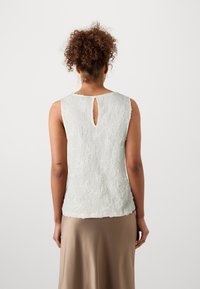 Top sin mangas de textura ligera en verde pálido con patrones florales y una abertura en forma de llave en la parte posterior. Combinado con una falda lisa en color beige.
