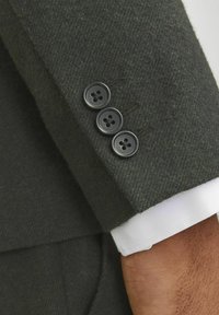 Tmavozelená vlnená sako má rukáv s tromi čiernymi gombíkmi s štyrmi otvormi; pod ním je vidieť biely manžetový rukáv.