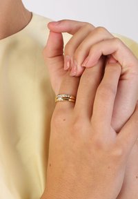 Goldring-Set mit zwei Bändern, wobei eines mit klaren Edelsteinen verziert ist. Hände zeigen glatte Haut mit leichtem Nagellack. Der Hintergrund ist sanft gelb.