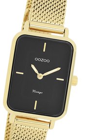 Montre avec bracelet en mailles dorées et cadran noir rectangulaire. Présente des index horaires dorés, des aiguilles simples, et le texte "Vintage" en bas.