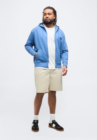 Άνδρας με κοτσίδες, που φοράει μπλε φούτερ με φερμουάρ, λευκό t-shirt, μπεζ σορτς, λευκές κάλτσες και μαύρα αθλητικά παπούτσια, στέκεται μπροστά σε λευκό φόντο.