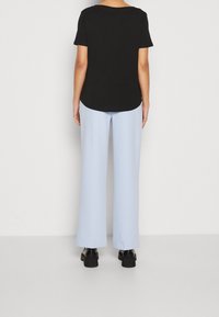 Osoba stojąca tyłem, ubrana w czarną koszulę z krótkim rękawem, jasnoniebieskie szerokie spodnie i czarne masywne buty, na jednolitym tle.