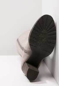 Sämjig knähöga stövlar i ruggat läder med en texturerad svart gummisula och klotformad klack, vilande på sidan mot en vit bakgrund och vägg.