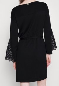 Černé šaty s ribbed horní částí, krajkovými zvonovými rukávy, páskem v oblasti pasu a malým zlatým akcentem na výstřihu.