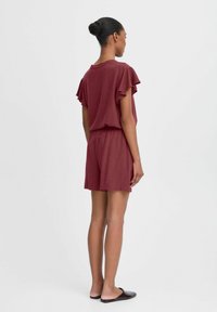 Une combinaison courte bordeaux avec des manches volantes, une taille élastique et un tissu doux. Texture lisse ; coupe décontractée idéale pour le confort et le mouvement.